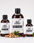 Three amber glass bottles of The Skin Science Cherry Kernel Oil in 200ml, 500ml, and 150ml sizes, arranged with shelled almonds, green leaves, and a cherry on a white background.