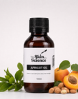 A 100ml bottle of Apricot Oil by The Skin Co. Science, presented in an amber glass bottle with a black cap and a white label, styled with fresh apricots, a halved apricot, green leaves, and almonds on a clean white background.
