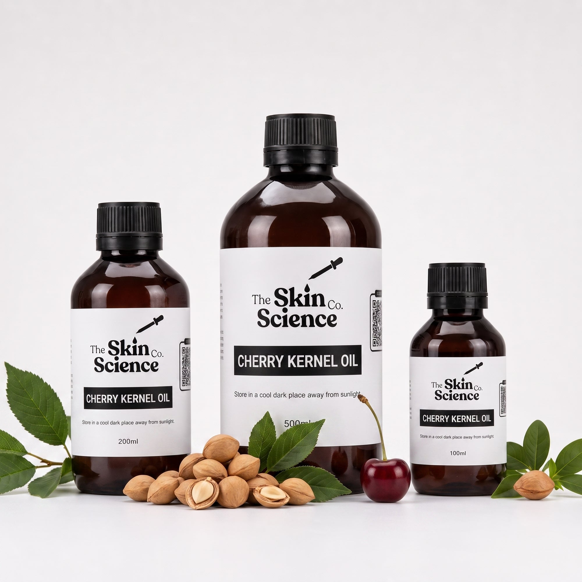 Three amber glass bottles of The Skin Science Cherry Kernel Oil in 200ml, 500ml, and 150ml sizes, arranged with shelled almonds, green leaves, and a cherry on a white background.