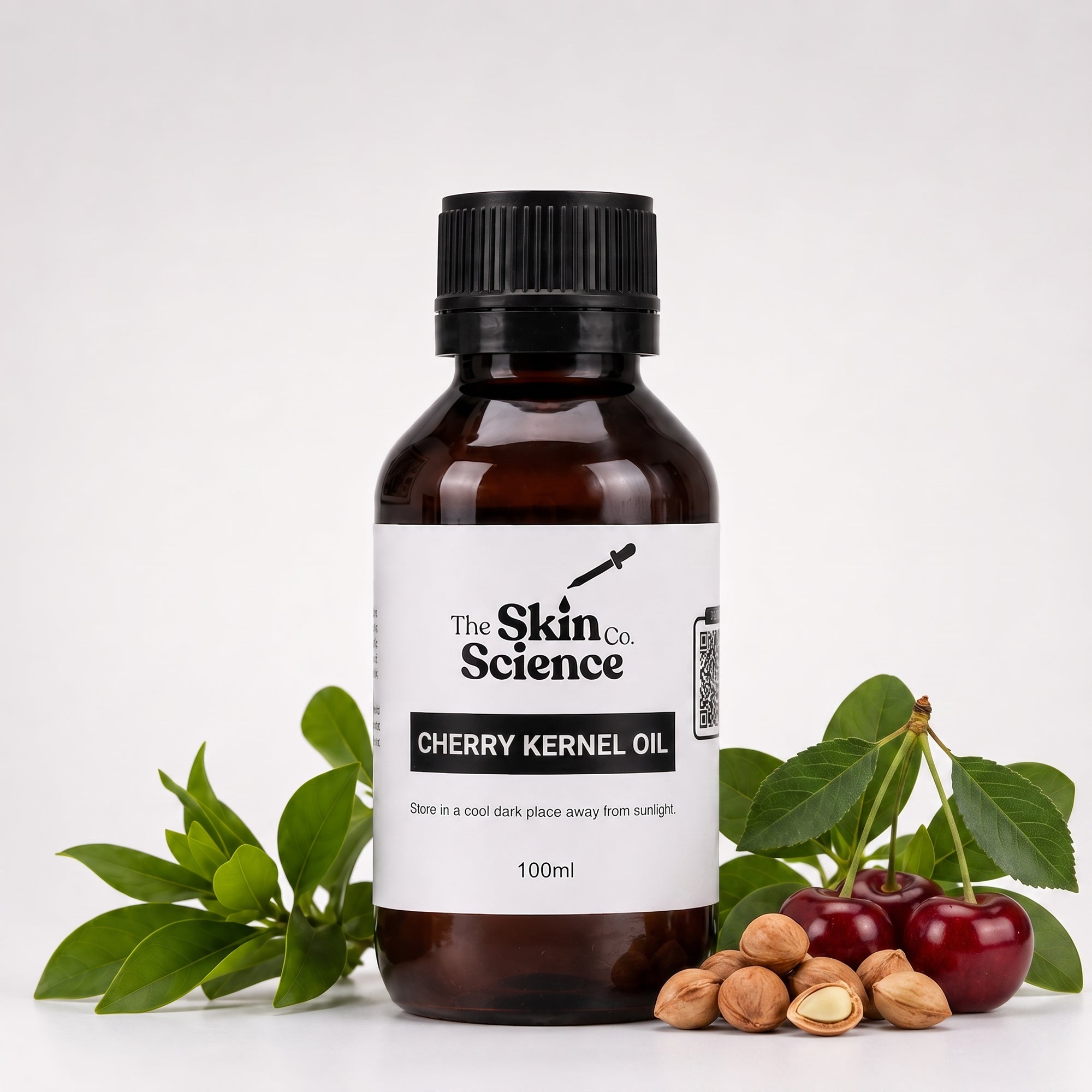 A 100ml bottle of Cherry Kernel Oil by The Skin Co. Science, in an amber glass bottle with a black cap and a white label, styled with fresh cherry leaves, cherries, and whole nuts on a white background.